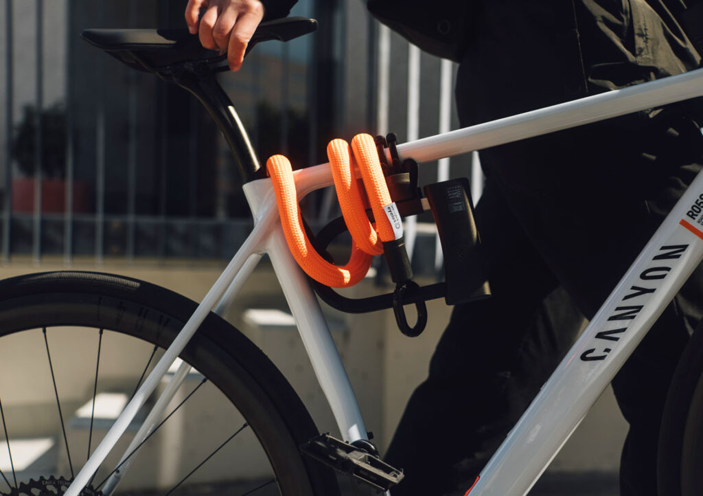 D-lock Bügelschloss mit Halterung am weißen Canyon-Fahrradrahmen transportiert, mit tex–lock eyelet-Textilseil in neon orange um's Oberrohr gewickelt